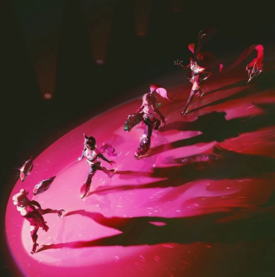 A high-angle view of several dancers performing on a glossy pink circular stage under dramatic red and pink lighting, with long shadows stretching across the dark background.