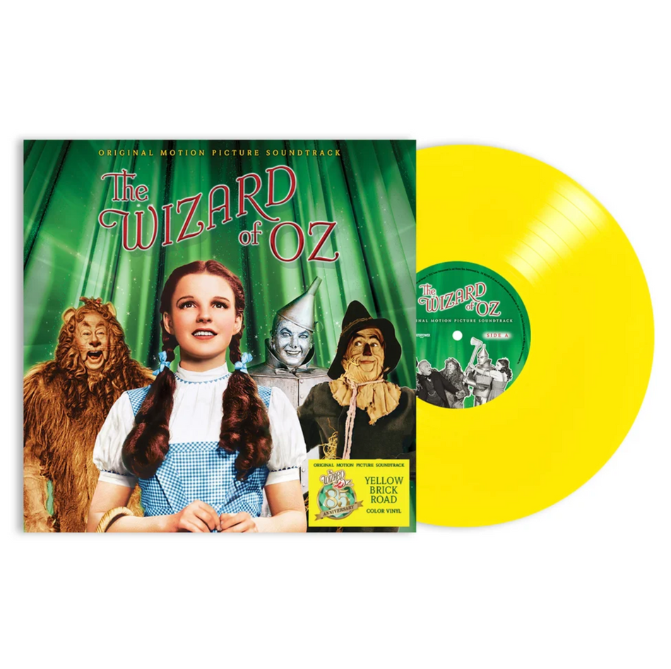 The Wizard of Oz motion picture soundtrack vinyl jacket art featuring Dorothy, the Cowardly Lion, the Tin Man, and the Scarecrow all against a shimmering green background. A sticker in the bottom right mentions the variant being yellow brick road yellow. The disc sticking out the side of the jacket is an opaque bright yellow with a green center label, also featuring the same 4 characters as the front but in black and white.
